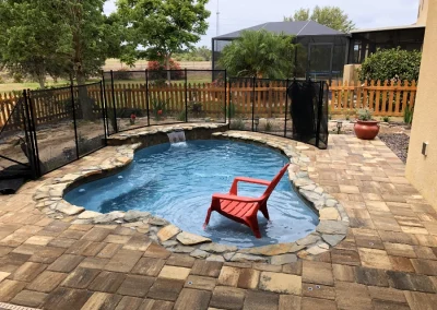 A small backyard pool with a stone border features a distinctive red chair in the water, all safely enclosed by a fence. The area, enhanced by expert pool construction, is complemented by a brick patio and lush garden with plants and trees in the background. Flamingo Pools, Punta Gorda FL