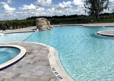 Outdoor swimming pool with clear blue water and a stunning rock feature under a partly cloudy sky. Poolside tiles, expertly laid during the recent pool construction, boast depth markings visible around the edges, enhancing both safety and style. Flamingo Pools, Punta Gorda FL