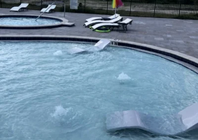 A circular shallow pool with submerged lounge chairs and a red-and-yellow flag on the deck. In the background, a freshly remodeled small pool, more lounge chairs, a fence, and a cloudy sky create an inviting oasis. Flamingo Pools, Punta Gorda FL