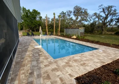 Rectangular outdoor swimming pool surrounded by stone tiles, adjacent to a house, showcases a recent pool remodel. Trees and grass are visible in the background. Flamingo Pools, Punta Gorda FL