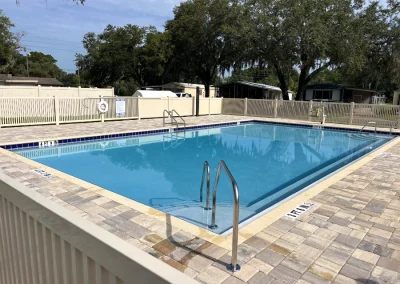 A rectangular outdoor swimming pool with clear water, recently refreshed by a top-notch pool remodel, is surrounded by a paved area and a white fence. The area is empty, with trees and buildings in the background. Flamingo Pools, Punta Gorda FL