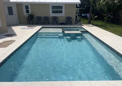 A rectangular swimming pool, recently revamped through a skilled pool remodel, features a small attached spa. It's surrounded by a concrete deck with several gray chairs, sitting adjacent to a beige house, offering both style and comfort. Flamingo Pools, Punta Gorda FL