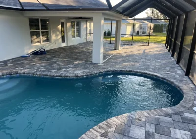 The outdoor swimming pool with blue water sits gracefully on a brick patio, covered by a screened enclosure. Adjacent to the white house with its shaded porch, this beautifully crafted pool is a testament to exceptional pool construction, with a visible lawn in the background. Flamingo Pools, Punta Gorda FL