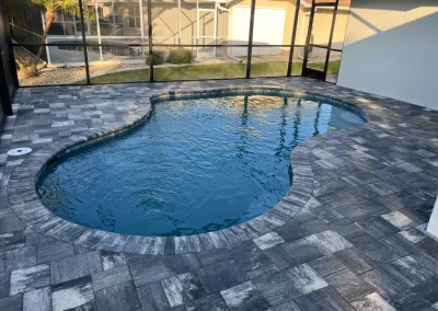 An enclosed backyard pool with a stone tile patio, curved edges, and clear blue water, surrounded by a screened structure, invites relaxation. It's the perfect setting to consider a pool remodel for that ultimate personal oasis. Flamingo Pools, Punta Gorda FL