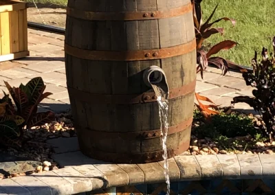 A wooden barrel fountain flows into a pool, expertly crafted through skilled pool construction, set on a stone patio with nearby plants, under a sunny sky. Flamingo Pools, Punta Gorda FL