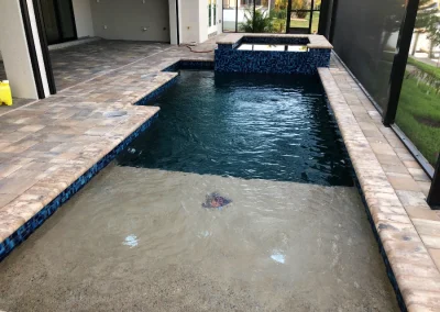 A beautifully crafted covered patio features a small rectangular pool with expert pool construction and an adjoining spa. The area is elegantly paved with stone tiles, while a landscaped garden is visible in the background, completing this serene oasis. Flamingo Pools, Punta Gorda FL