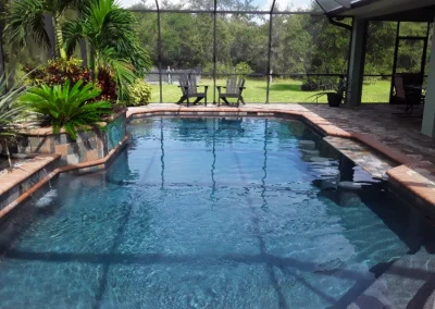 A rectangular backyard pool with a screened enclosure is nestled among lush foliage. In the background, a small patio features two chairs and a table—an inviting spot to relax after your dream pool remodel. Flamingo Pools, Punta Gorda FL