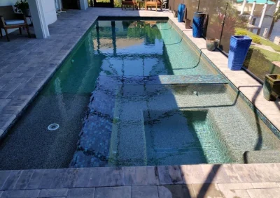 This beautifully remodeled rectangular pool features decorative tiles, steps, and a submerged lounge ledge. It's surrounded by a stone patio with blue planters, offering a partial view of nearby furniture and lush greenery—perfect for relaxed outdoor living. Flamingo Pools, Punta Gorda FL