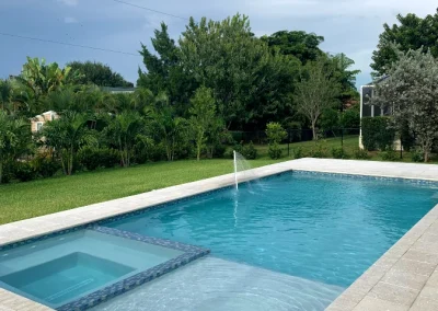 A rectangular swimming pool with a hot tub is set amidst a stone tile deck, evoking tranquility. Lush green trees and grass surround the area under a cloudy sky, offering the perfect escape. Consider our pool remodel services to enhance your backyard oasis even further. Flamingo Pools, Punta Gorda FL