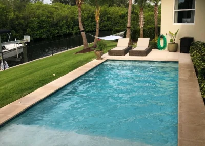 A rectangular outdoor swimming pool, impeccably maintained by a top-notch pool service, is bordered by a patio, three lounge chairs, and palm trees, with a view of a canal and greenery in the background. Flamingo Pools, Punta Gorda FL