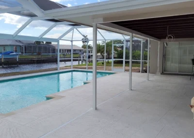 A covered patio with a swimming pool, freshly treated by a reliable pool service, overlooks a serene canal. A dog strolls on the patio as houses with screened porches are visible across the canal. Flamingo Pools, Punta Gorda FL