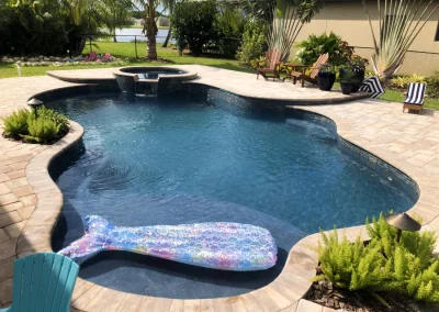 A beautifully remodeled backyard pool with a stone patio, surrounded by lush plants. A whale-shaped inflatable float gently bobs in the water, while striped lounge chairs invite relaxation on the newly revamped patio. Flamingo Pools, Punta Gorda FL