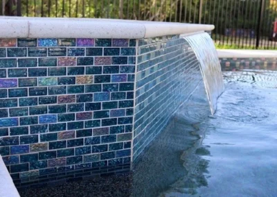 A close-up of a swimming pool's edge showcases a vibrant mosaic tile design, and a waterfall feature elegantly cascades into the pool. The scene, artfully crafted through expert pool construction, is framed by a metal fence with trees in the background. Flamingo Pools, Punta Gorda FL