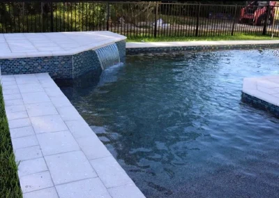 This stunning rectangular swimming pool, a result of recent pool construction, features a small waterfall. It's beautifully enclosed by a black metal fence and surrounded by grey tile decking and lush grass. Flamingo Pools, Punta Gorda FL