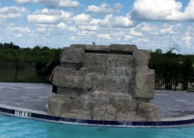 A rock waterfall feature enhances this swimming pool with its clear blue water. In the background, a fence and trees complement a sky of scattered clouds, showcasing the elegance of recent pool construction. Flamingo Pools, Punta Gorda FL