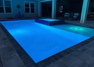 The rectangular swimming pool and hot tub, crafted with precision by an expert pool construction team, are illuminated with blue lights at night. They are surrounded by a tiled patio and stylish outdoor furniture. Flamingo Pools, Punta Gorda FL