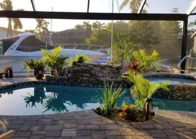 Enclosed pool area with a stone border, surrounded by plants. A yacht is visible through the screened enclosure, and palm trees are in the background. The space boasts recent pool remodel touches enhancing its beauty. Flamingo Pools, Punta Gorda FL