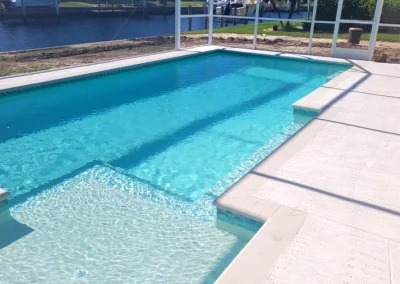 Rectangular swimming pool with clear blue water, surrounded by a white concrete deck and enclosed by a screen. Recently enhanced through expert pool remodel, it features a small step-in area and waterway visible in the background. Flamingo Pools, Punta Gorda FL