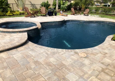 A luxurious backyard pool with an attached circular hot tub, expertly crafted by top-notch pool construction professionals. Surrounded by paver tiles and several wooden lounge chairs, it offers a serene escape with plants and a fence providing privacy in the background. Flamingo Pools, Punta Gorda FL