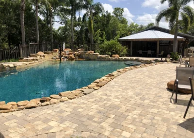 This outdoor pool, showcasing expert pool construction, features a rock border and an adjacent patio, all embraced by towering trees. In the background, a charming wooden structure with a metal roof completes this tranquil setting. Flamingo Pools, Punta Gorda FL